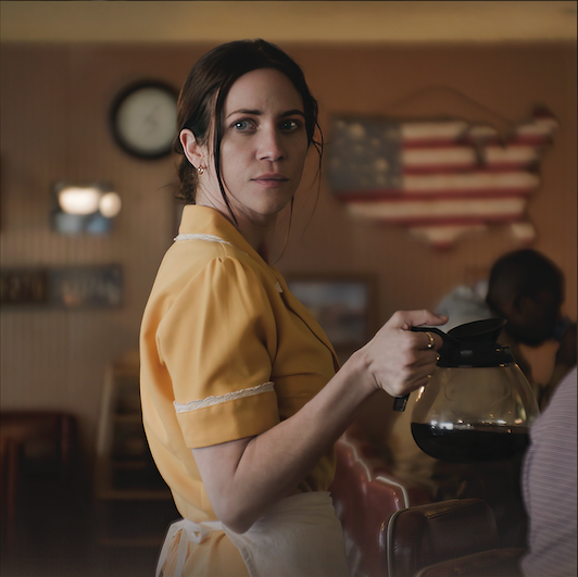 Waitress wearing a yellow uniform stands in diner holding a pot of coffee.