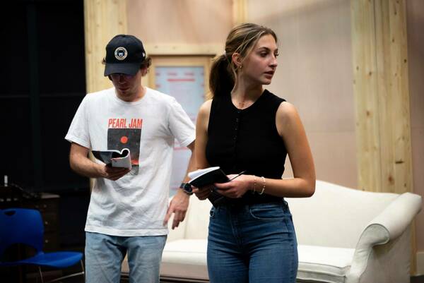 Two individuals stand in a room, engaged with books. The person on the left wears a white Pearl Jam t-shirt and a navy baseball cap with a gold circular logo. They hold an open book. The person on the right, wearing a black sleeveless top and jeans, holds a smaller book and looks to the side.