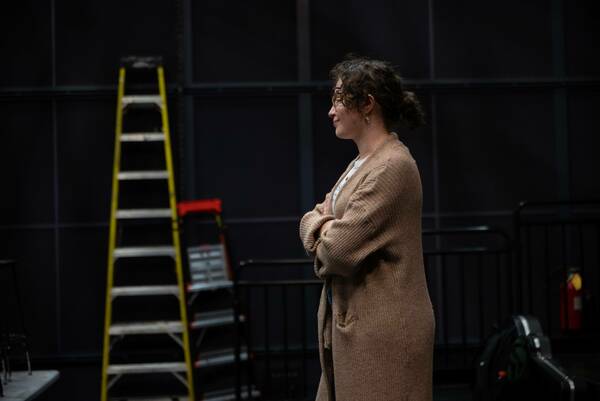 A person with shoulder-length curly hair, wearing glasses and a long, light brown cardigan, stands with their arms crossed, looking to the right. A yellow and silver ladder is slightly blurred in the background to the left. Black sound dampening panels line the wall behind them.