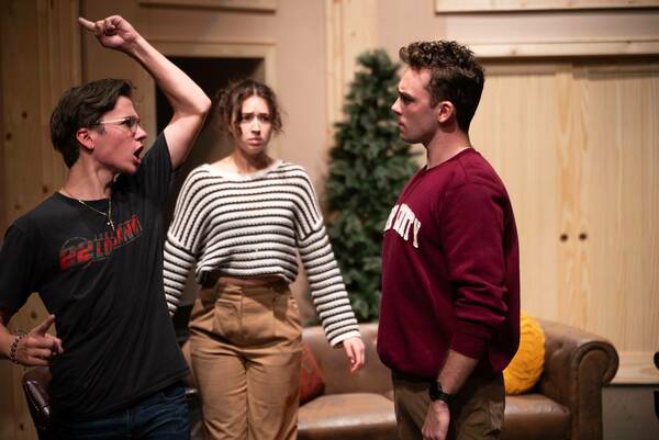 Three students perform in a play on a stage with a partial set, including a couch. On the left, a person with their arm raised high looks toward center stage with their mouth open in apparent shock. In the center, a student wearing a black and white striped sweater looks concerned. On the right, a student wearing a maroon sweatshirt looks calmly to the left.