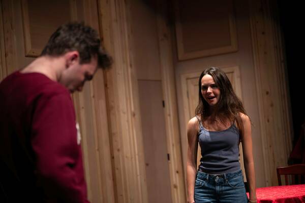Two actors on a sparsely decorated stage with wood-paneled walls. The woman, wearing jeans and a tank top, addresses the man, who is slightly out of focus and wearing a maroon shirt.