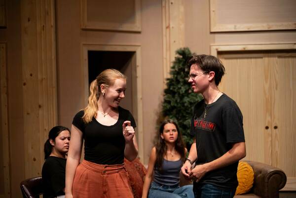 On a stage with muted lighting and a backdrop resembling a living room, two student actors engage in a scene. A blonde female student, wearing a black top and burnt orange pants, gestures with her right hand while looking at a male student. He wears glasses, a black t-shirt with a Joey Lugano graphic, and jeans. In the background, two other students observe the scene.