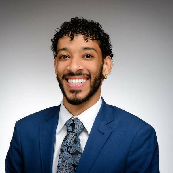 A young man with dark curly hair and a beard smiles broadly. He wears a blue suit, white shirt, and a dark paisley tie. He has a nose piercing and gold hoop earrings.