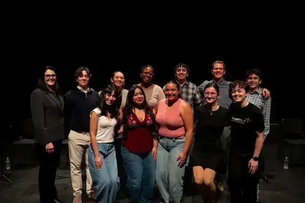 A diverse group of 12 smiling people stand on a stage with a black background. One person wears a "DEBARTOLO ARTS CENTER" shirt.