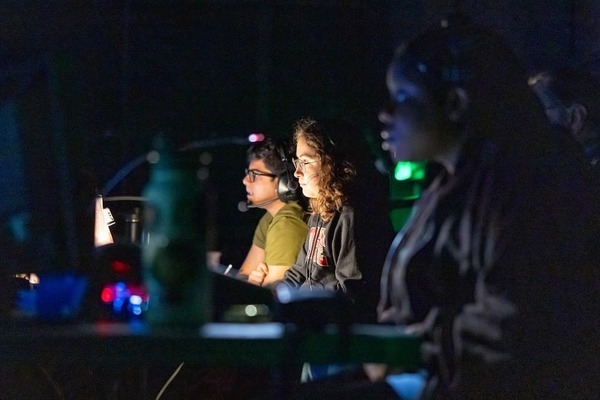 Male and female student crew members sit at a desk in a dark room watching a rehearsal for a play