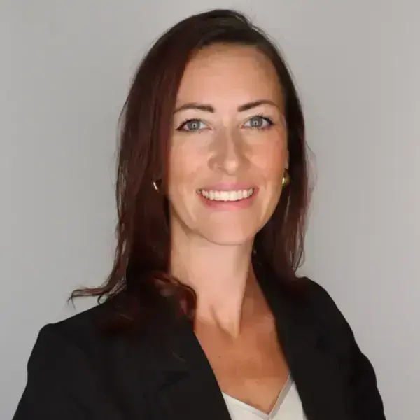 A woman with reddish-brown hair and blue eyes smiles, wearing a black blazer over a light top and gold hoop earrings.