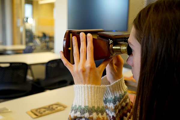 Young woman with brown hair looks intently into a vintage wooden stereoscope with a brass eyepiece, wearing a patterned sweater.